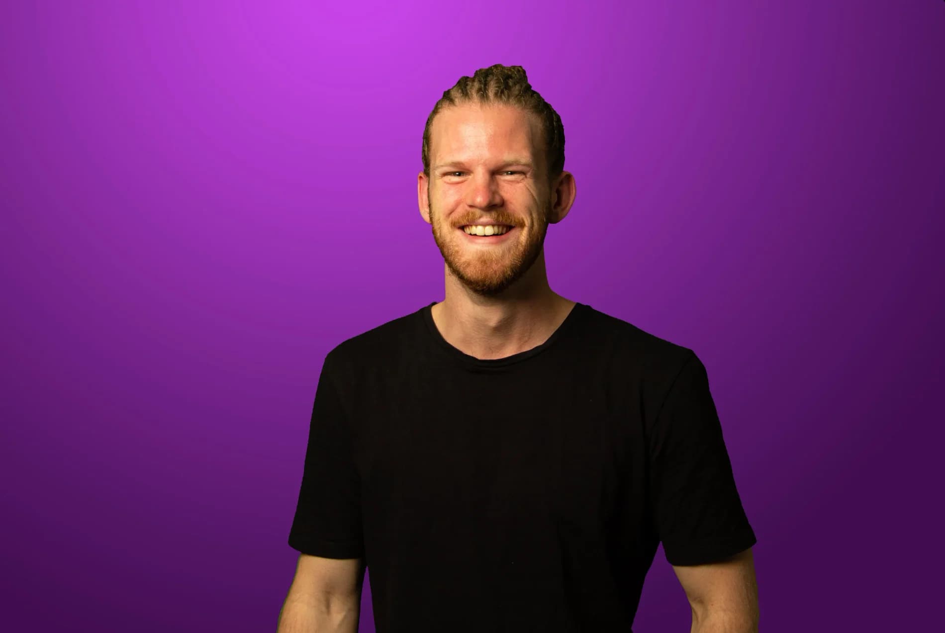 Emanuel Hansson ler brett i svart t-shirt och uppsatta flätor mot lila bakgrund