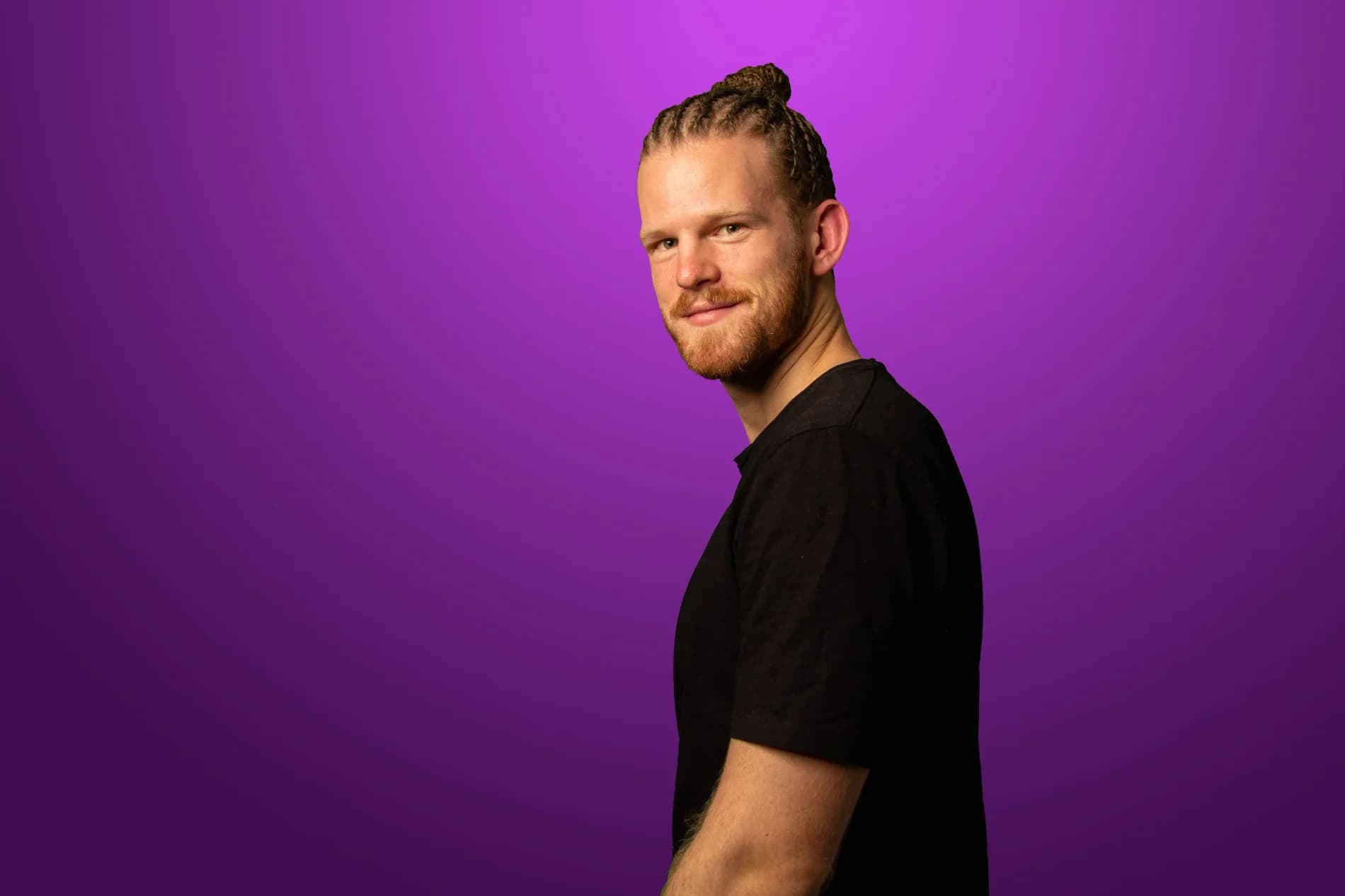 Emanuel Hansson i profil vänder huvudet mot kameran och ler, flätorna i knut, mot lila bakgrund