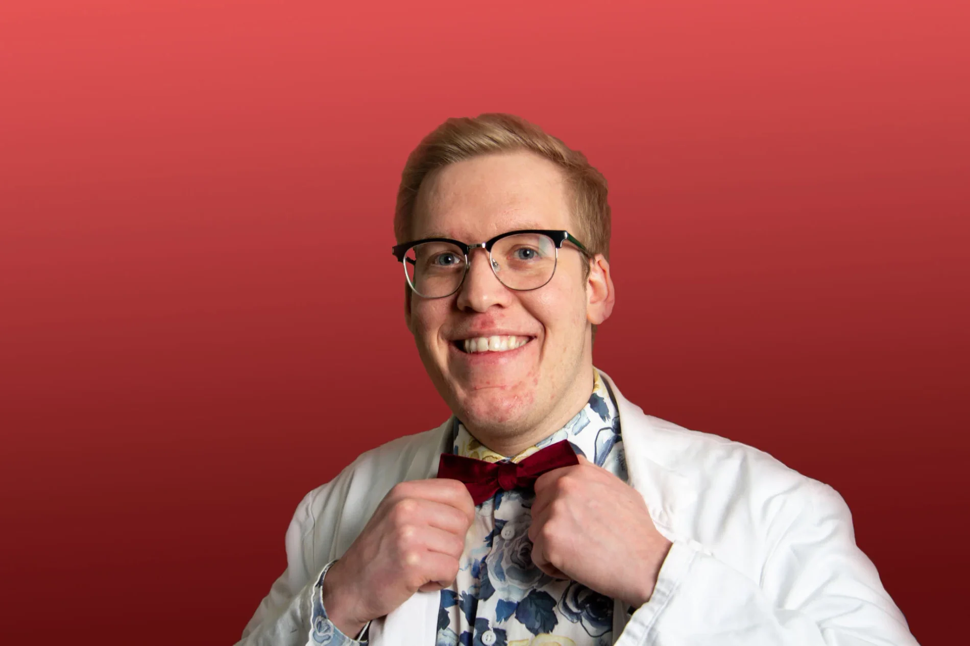 Freddy Grip rättar till sin vinröda fluga och ler stort, i vit labbrock mot röd bakgrund