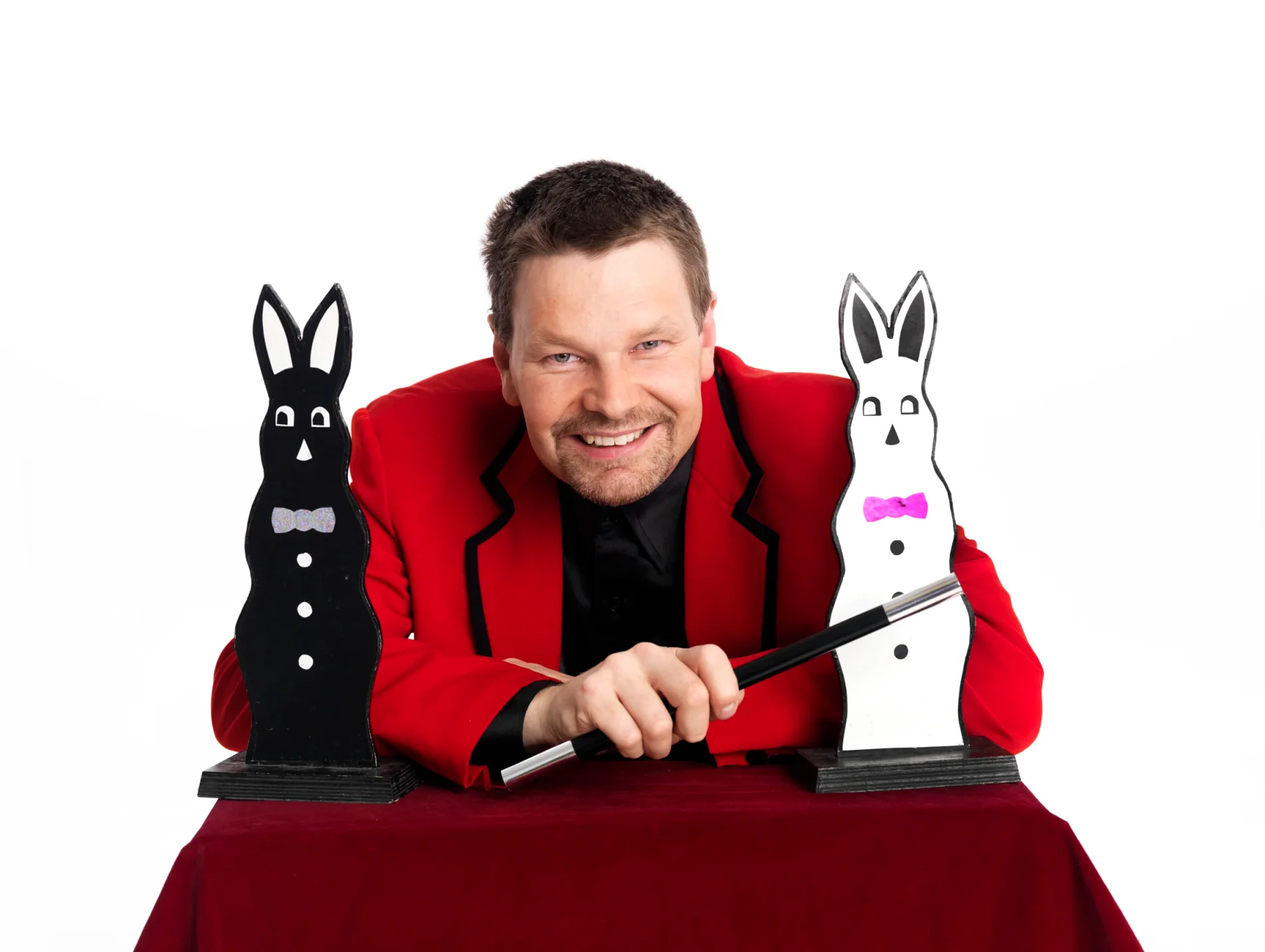 Mr. Goran i röd kavaj håller ett trollspö mellan två kaninfigurer på ett sammetsklätt bord