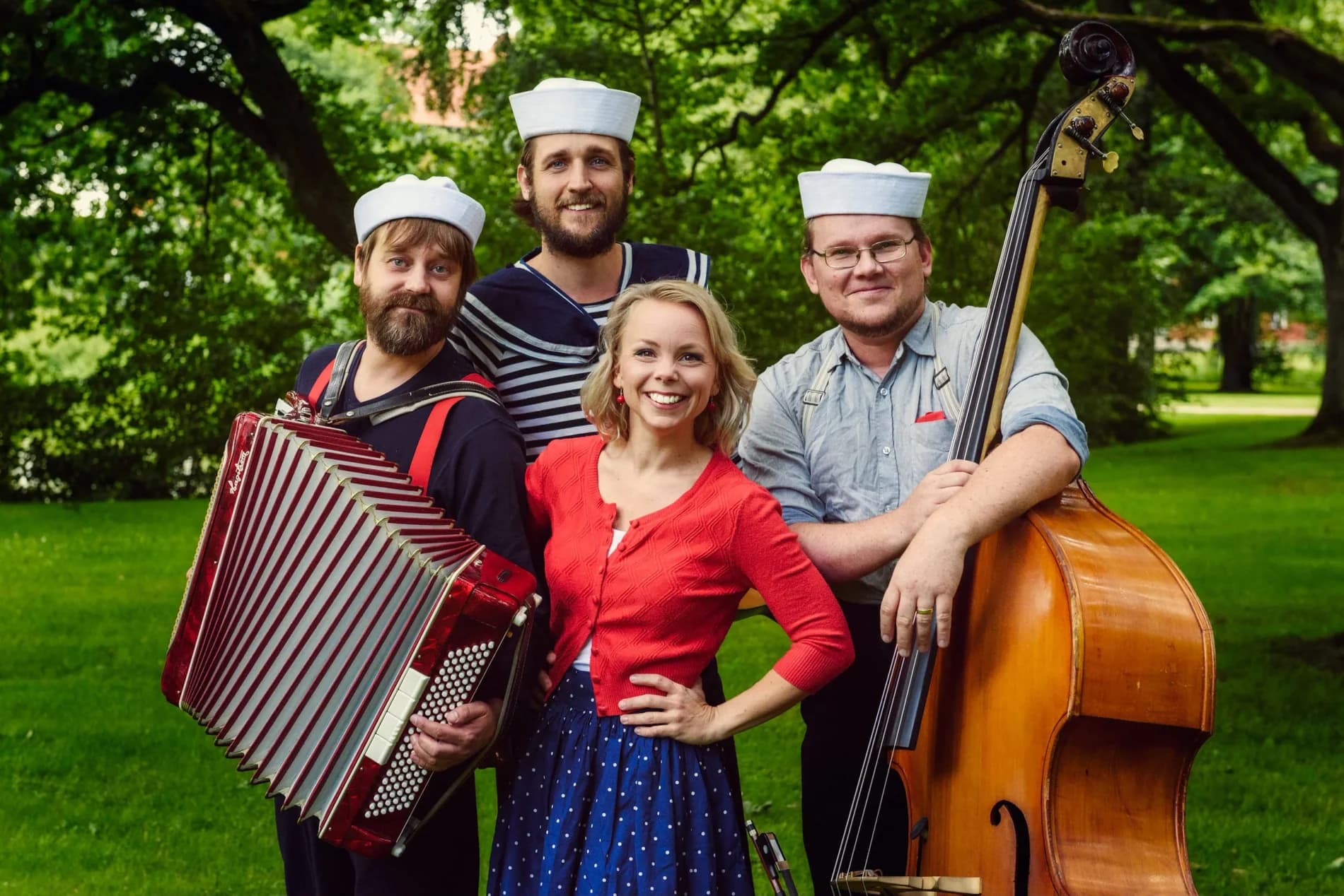 Nina Hjelmkvist i röd kofta med sina tre musiker i sjömansmössor, dragspel och kontrabas i en park