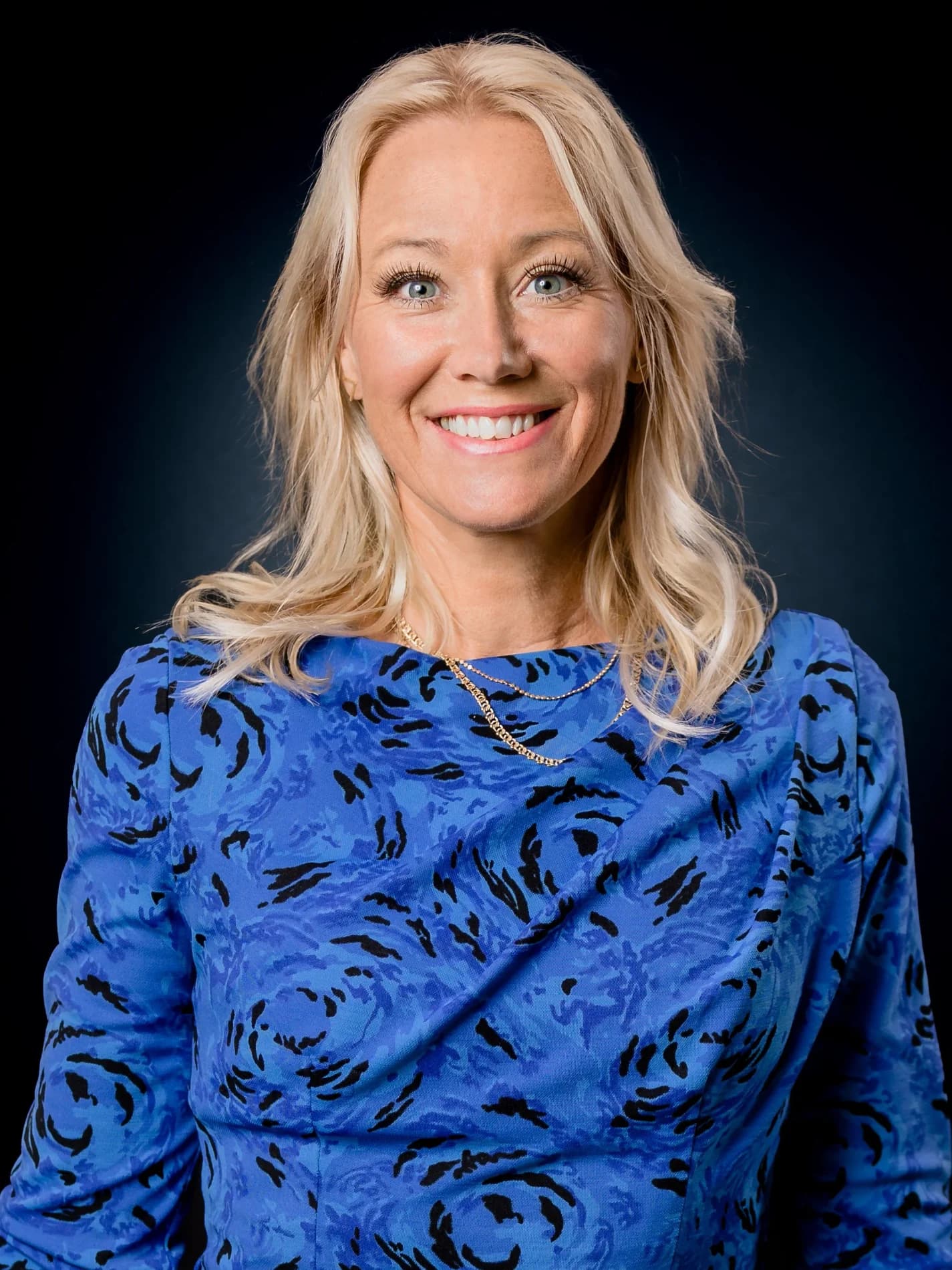 Sara Edwardsson ler stort i blåmönstrad klänning och guldhalsband mot mörk bakgrund