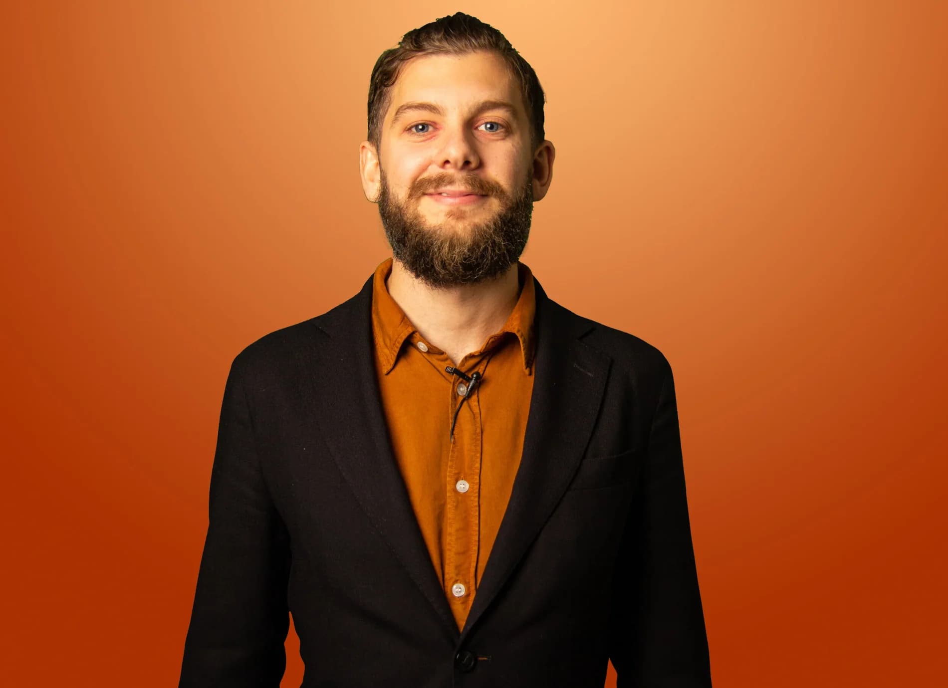 Simon Söderqvist i svart kavaj och orange skjorta ler mot kameran framför orange fond